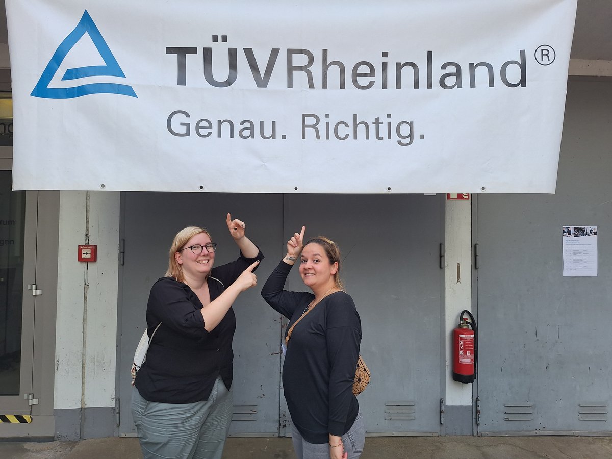 Zwei Frauen zeigen auf Schild mit Tüv Rheinland Logo